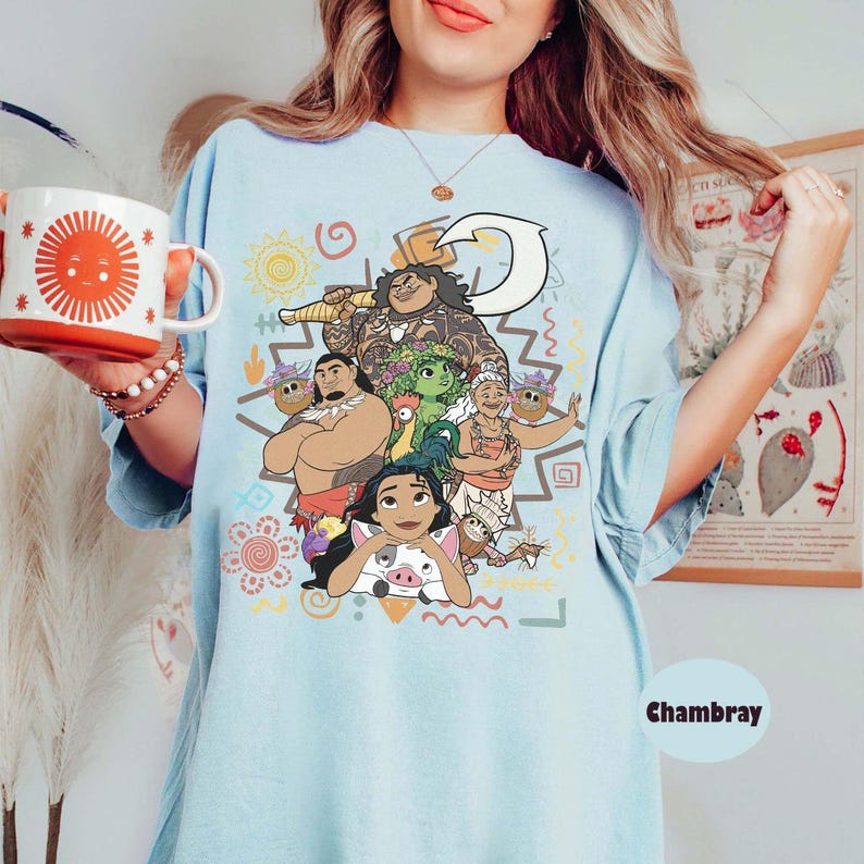 May include: A light blue t-shirt featuring a colorful graphic print of characters from the movie Moana, including Moana, Maui, Pua, Heihei, and Gramma Tala. The shirt is paired with a white mug with a large orange sun design on the front.