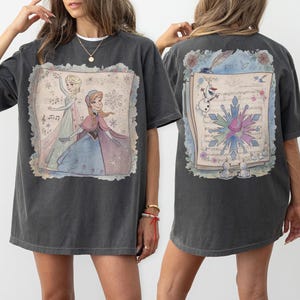 Könnte beinhalten: Dunkelgraues T-Shirt mit einer farbenfrohen Illustration von Elsa und Anna aus Frozen, Noten und Schneeflocken. Auf der Rückseite befindet sich ein passendes Design mit Olaf und einer Schneeflocke. Ein lustiges, lässiges T-Shirt.