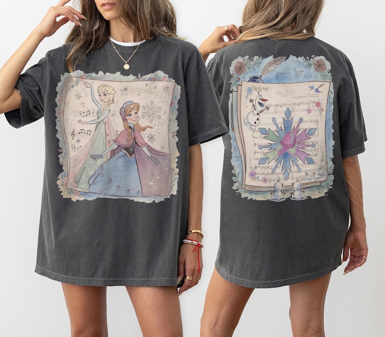 Pode incluir: T-shirt cinzento escuro com uma ilustra&ccedil;&atilde;o colorida de Elsa e Anna de Frozen, com notas musicais e flocos de neve. A parte de tr&aacute;s da camisa tem um design a condizer com Olaf e um floco de neve. Uma t-shirt divertida e casual.
