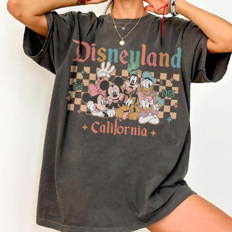 May include: Dark gray t-shirt featuring the word "Disneyland" in colorful text above a graphic of Mickey, Minnie, Goofy, Donald, and Pluto. The shirt also includes the text "Est. 1955 California" with a checkered background.