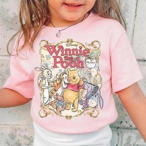 May include: Pink short-sleeved t-shirt featuring a graphic of Winnie the Pooh and friends. The design includes Winnie the Pooh, Tigger, Eeyore, Piglet, and Rabbit, with the words "Winnie the Pooh" in pink script.