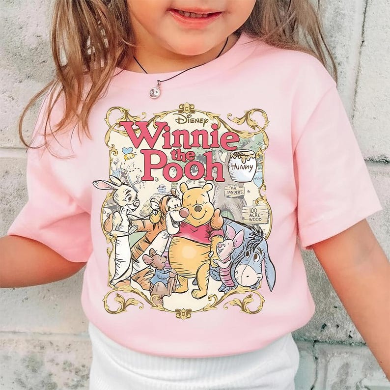 May include: Pink short-sleeved t-shirt featuring a graphic of Winnie the Pooh and friends. The design includes Winnie the Pooh, Tigger, Eeyore, Piglet, and Rabbit, with the words "Winnie the Pooh" in pink script.