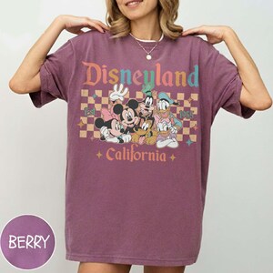 May include: Berry-colored t-shirt featuring a Disneyland graphic with Mickey Mouse, Minnie Mouse, Goofy, Donald Duck, and Daisy Duck. The design includes the text "Disneyland California Est. 1955" in a retro style. The shirt has short sleeves and a relaxed fit.