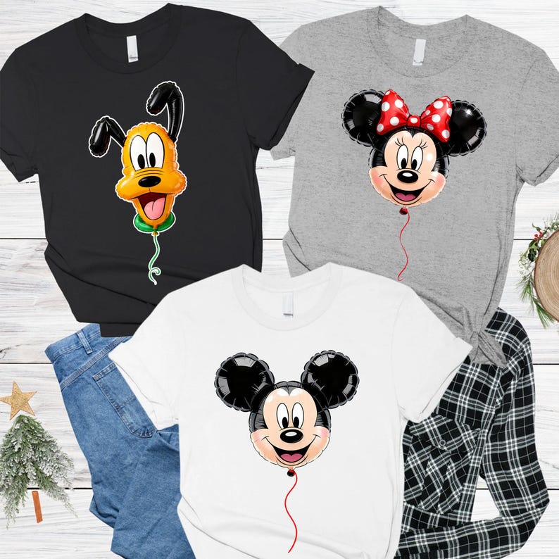 May include: A black t-shirt with a Pluto balloon graphic, a grey t-shirt with a Minnie Mouse balloon graphic featuring a red and white polka dot bow, and a white t-shirt with a Mickey Mouse balloon graphic, all featuring short sleeves and crew necks.