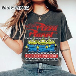 May include: A pepper-colored t-shirt featuring a red Pizza Planet logo, three green three-eyed aliens in blue uniforms, and the text Food & Fun Space Port Serving your local star cluster. The design includes a black and white checkered border.
