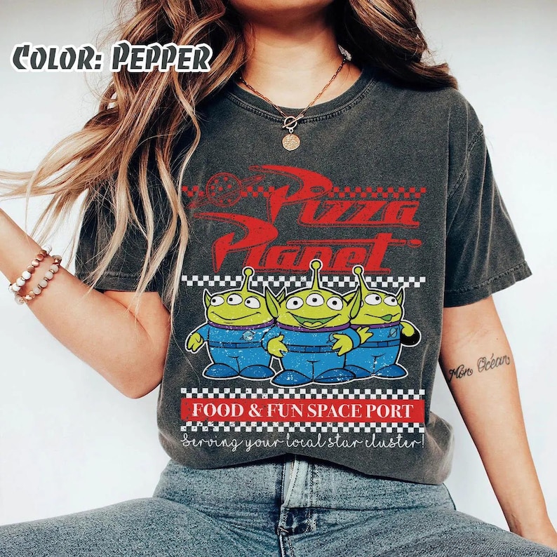 May include: A pepper-colored t-shirt featuring a red Pizza Planet logo, three green three-eyed aliens in blue uniforms, and the text Food & Fun Space Port Serving your local star cluster. The design includes a black and white checkered border.