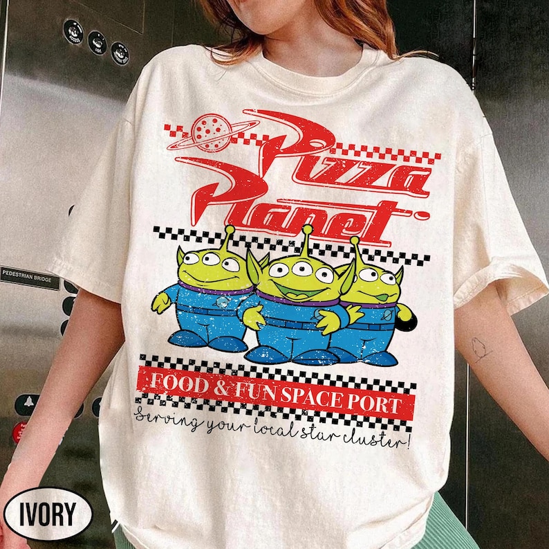 May include: An ivory t-shirt featuring a red and black graphic print of three green three-eyed aliens in blue uniforms. The text reads Pizza Planet, Food & Fun Space Port, and Serving your local star cluster, with a checkered pattern border.