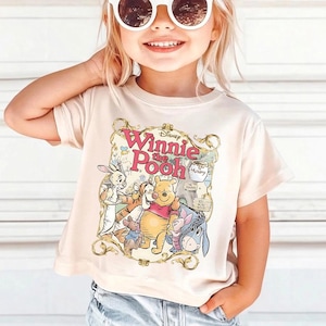 May include: A beige t-shirt featuring a colorful graphic of Winnie the Pooh and friends. The design includes the text "Disney Winnie the Pooh" in a decorative frame. The shirt is short-sleeved and has a crew neck.