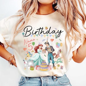 Pode incluir: Camiseta creme com um design de anivers&aacute;rio colorido. O gr&aacute;fico inclui a palavra "Birthday", o n&uacute;mero "5" e imagens de uma princesa, um pr&iacute;ncipe, um bolo e bal&otilde;es. O design &eacute; festivo e comemorativo.