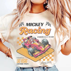May include: Cream-colored sweatshirt featuring a graphic of Mickey Mouse in a Formula 1 race car. The design includes the words "MICKEY Racing" and "Formula 1 Las Vegas Grand Prix". The car is pink and yellow with the number 28.