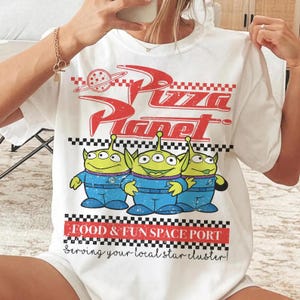 May include: A white t-shirt featuring a red Pizza Planet logo with a planet graphic, a black and white checkered pattern, and three green three-eyed aliens in blue space suits. Below is the text FOOD & FUN SPACE PORT and Serving your local star cluster.