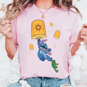 May include: A light pink t-shirt featuring a graphic of a blue alien character holding a large yellow lantern with a sun design. A small green lizard character is positioned below the alien, surrounded by several smaller yellow lanterns and stars.
