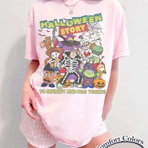May include: A pale pink t-shirt with a Halloween-themed graphic featuring cartoon characters and the text "HALLOWEEN STORY" and "TO INFINITY AND BOO YOUND!". The shirt is a Comfort Colors Blossom.
