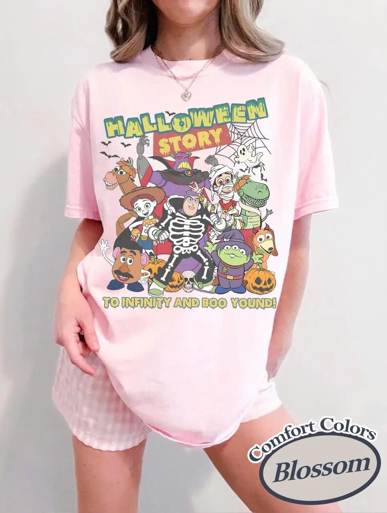 May include: A pale pink t-shirt with a Halloween-themed graphic featuring cartoon characters and the text "HALLOWEEN STORY" and "TO INFINITY AND BOO YOUND!". The shirt is a Comfort Colors Blossom.