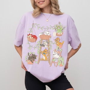 May include: A light purple t-shirt featuring a graphic of cartoon cats in hanging baskets and pots with green vines. The cats are in various colors, including orange, gray, and white. The shirt is a casual style.