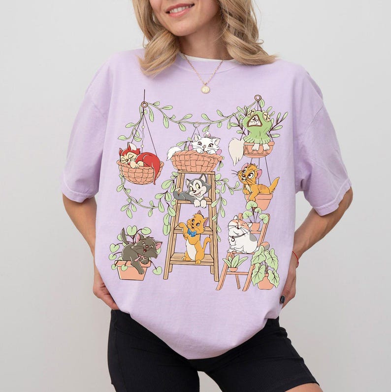 May include: A light purple t-shirt featuring a graphic of cartoon cats in hanging baskets and pots with green vines. The cats are in various colors, including orange, gray, and white. The shirt is a casual style.