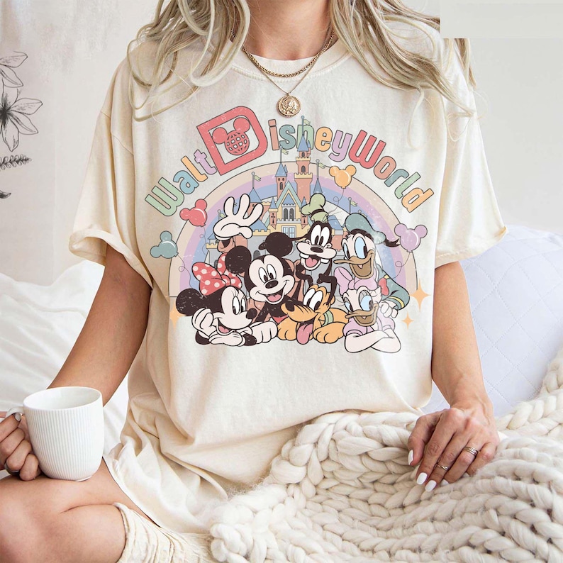 May include: Cream-colored t-shirt featuring a colorful graphic of Disney characters, including Mickey and Minnie Mouse, Goofy, Donald Duck, and Pluto, with the text "Walt Disney World" above a rainbow and castle.