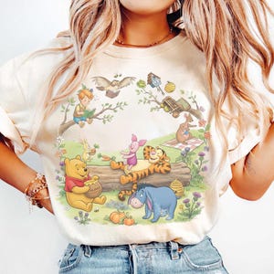 May include: Cream-colored t-shirt featuring a colorful illustration of Winnie the Pooh and friends. The design includes Pooh with a honey pot, Piglet, Tigger, Eeyore, and other characters in a whimsical outdoor scene. A charming graphic tee.