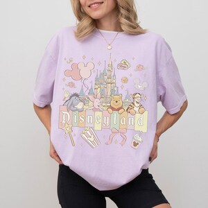 May include: A lilac t-shirt with a Disneyland graphic. The design features the Disneyland castle, Winnie the Pooh characters, and the word "Disneyland" in pastel shades. Other elements include balloons, popcorn, and a bow.