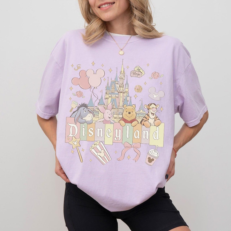 May include: A lilac t-shirt with a Disneyland graphic. The design features the Disneyland castle, Winnie the Pooh characters, and the word "Disneyland" in pastel shades. Other elements include balloons, popcorn, and a bow.