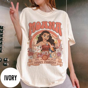 Op de afbeelding: Ivoor T-shirt met een afbeelding van Moana, het woord "Moana" in oranje retro lettertype. De afbeelding bevat illustraties van personages en bloemen, met de tekst "Est Disney Princess 2016" op een banner.