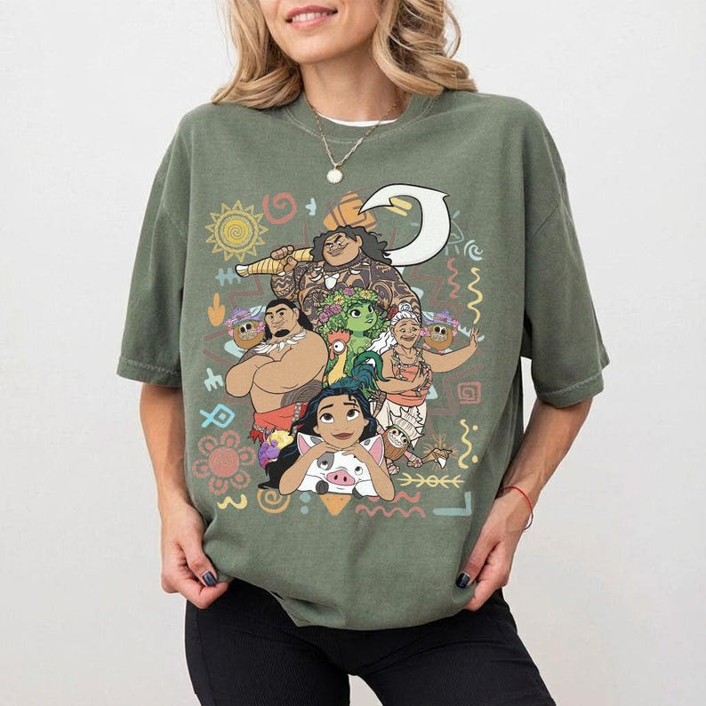 May include: An olive green short-sleeve t-shirt featuring a colorful graphic print of various animated characters, including a large figure holding a hook, a smaller central figure, and a pig, surrounded by stylized sun, wave, and tribal symbols.