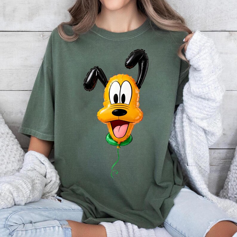 May include: A dark olive green short-sleeve t-shirt featuring a graphic print of a yellow dog character designed to look like a shiny foil balloon. The character has black ears, a wide open mouth with a pink tongue, and a green balloon knot.