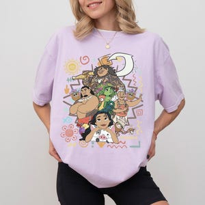 May include: A light purple short-sleeve t-shirt featuring a colorful graphic print of various animated characters, including a large figure holding a hook, a rooster, and a small pig. The design includes tribal-inspired patterns and symbols.