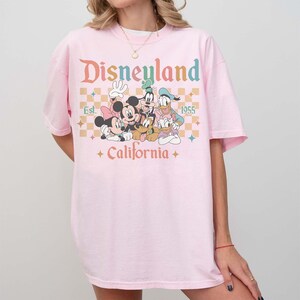 May include: A light pink t-shirt featuring the word "Disneyland" in a retro font, with a graphic of Mickey Mouse, Minnie Mouse, Donald Duck, Goofy, and Pluto. The shirt also includes the text "Est. 1955" and "California".