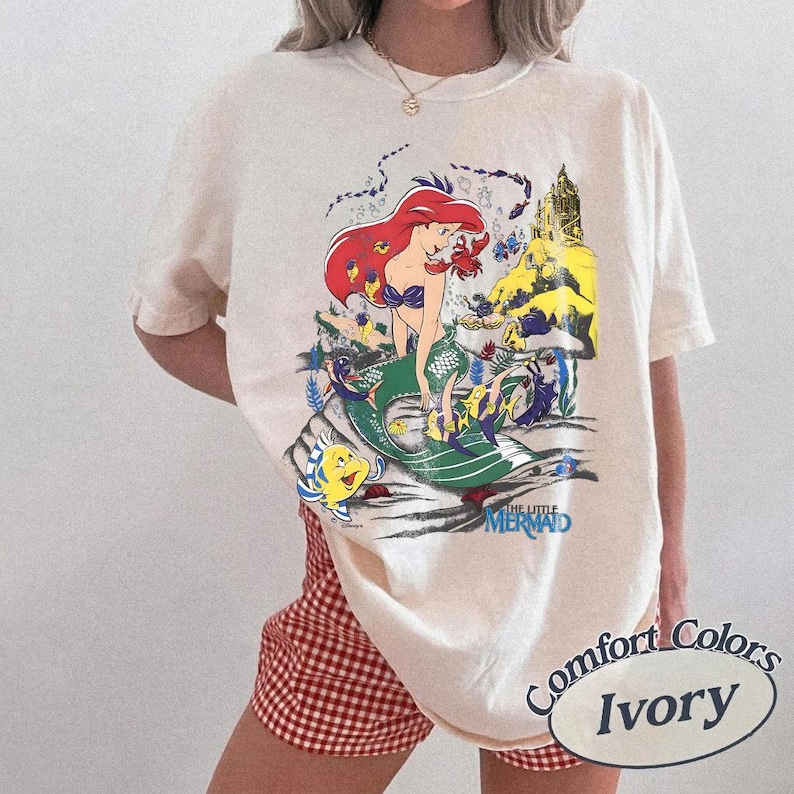 K&ouml;nnte beinhalten: Elfenbeinfarbenes T-Shirt mit einem Aufdruck von Arielle aus Arielle, die Meerjungfrau. Das Design zeigt Arielle mit roten Haaren und einem gr&uuml;nen Schwanz, zusammen mit anderen Charakteren und dem Text "THE LITTLE MERMAID". Das Shirt ist mit "Comfort Colors Ivory" gekennzeichnet.