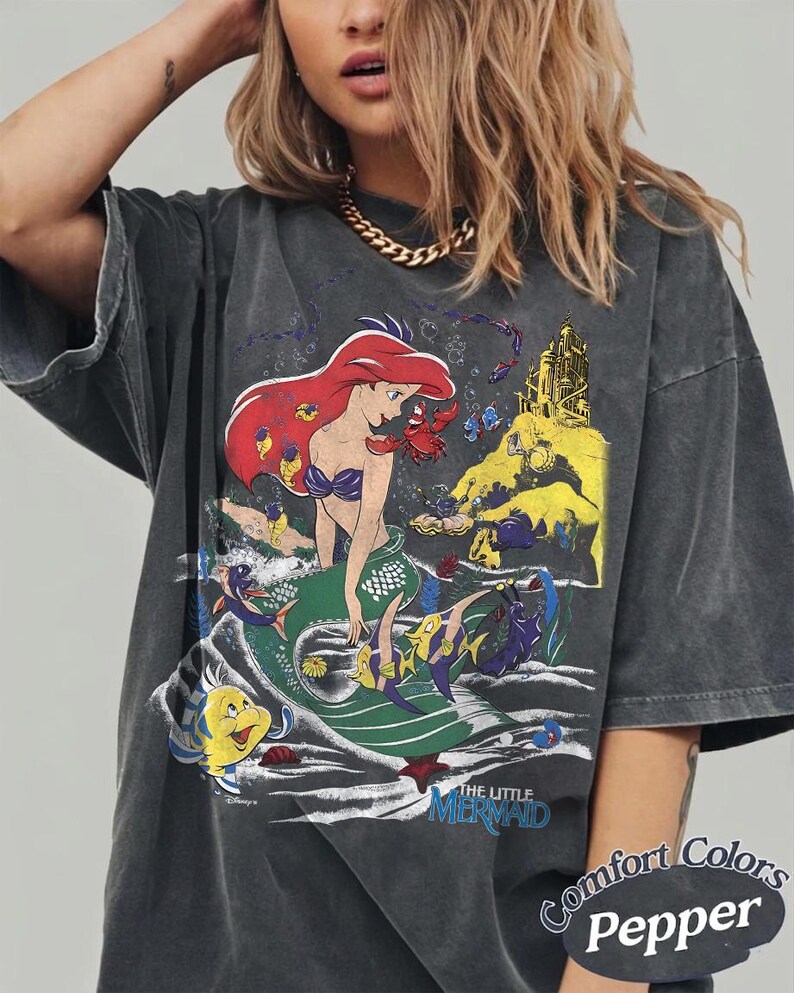 K&ouml;nnte beinhalten: Dunkelgraues T-Shirt mit einem Aufdruck von Arielle, der Meerjungfrau. Das Design zeigt Arielle, Fabius und ein Schloss. Der Text "THE LITTLE MERMAID" steht darunter. Das Shirt ist mit "Comfort Colors Pepper" gekennzeichnet.