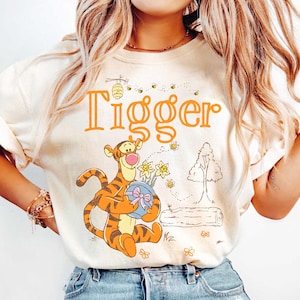 May include: Cream-colored t-shirt featuring the word "Tigger" in orange, with an illustration of Tigger holding a blue ball with a pink bow and yellow flowers. The design includes bees, a tree, and a log.