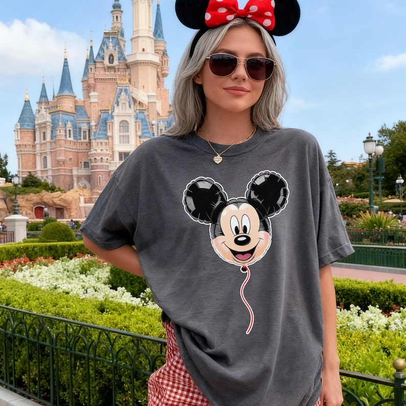 May include: A charcoal grey short-sleeve t-shirt featuring a graphic print of a Mickey Mouse-shaped balloon. The balloon is black with a light-coloured face, and a thin red string hangs down from the bottom of the graphic design on the front.