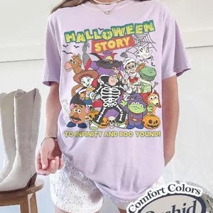 May include: A lilac t-shirt with a Halloween graphic featuring cartoon characters. The text "HALLOWEEN STORY" is at the top, with "TO INFINITY AND BOO YOUND!" below. The shirt is labeled "Comfort Colors" and "Orchid".