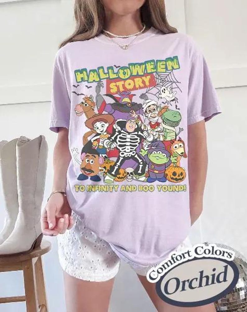 May include: A lilac t-shirt with a Halloween graphic featuring cartoon characters. The text "HALLOWEEN STORY" is at the top, with "TO INFINITY AND BOO YOUND!" below. The shirt is labeled "Comfort Colors" and "Orchid".
