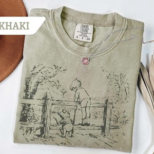 May include: Khaki-colored t-shirt featuring a black and white illustration of a boy and a bear looking over a wooden fence. The shirt has a crew neck and short sleeves. A silver necklace with a pink pendant is on the shirt.