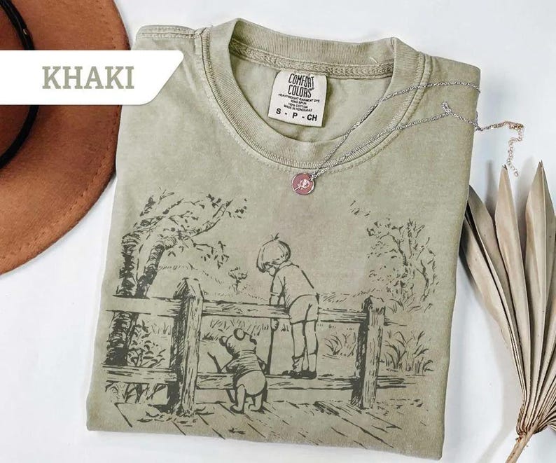 May include: Khaki-colored t-shirt featuring a black and white illustration of a boy and a bear looking over a wooden fence. The shirt has a crew neck and short sleeves. A silver necklace with a pink pendant is on the shirt.