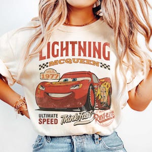 Puede incluir: Camiseta color crema con un dise&ntilde;o de Rayo McQueen de la pel&iacute;cula Cars. El dise&ntilde;o incluye el texto "LIGHTNING MCQUEEN" en rojo, junto con "Ultimate Speed" y "Think Fast". La camiseta tiene un aspecto vintage y desgastado.