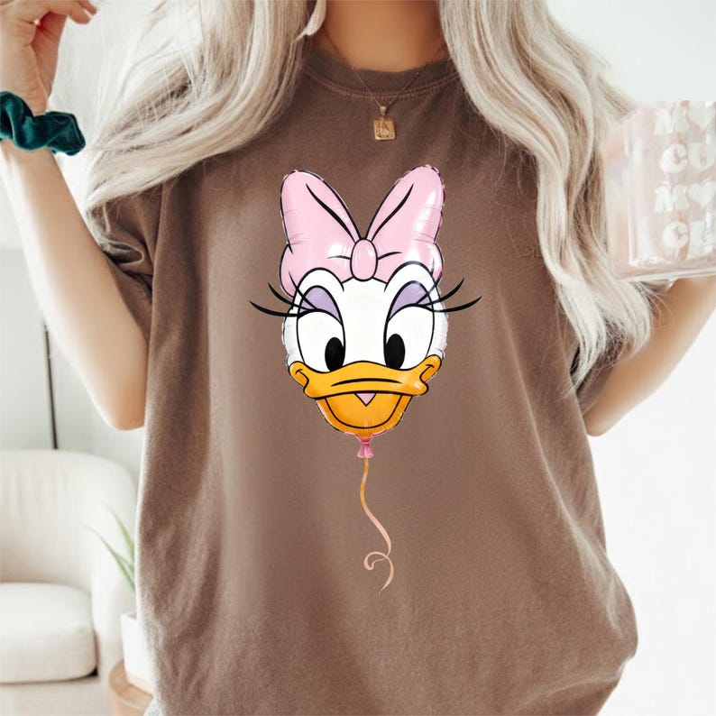 May include: A brown short-sleeve t-shirt featuring a graphic print of Daisy Duck's face designed as a pink and white balloon. The character has a pink bow, long black eyelashes, and an orange beak, with a thin pink string extending downward.
