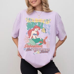 K&ouml;nnte beinhalten: Helllila T-Shirt mit einer Grafik von Arielle, der Meerjungfrau. Die Grafik zeigt Arielle, Fabius, Sebastian und den Text "Ocean Tour 1991". Das Shirt hat einen Rundhalsausschnitt und kurze &Auml;rmel.
