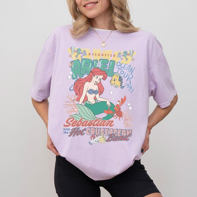 K&ouml;nnte beinhalten: Helllila T-Shirt mit einer Grafik von Arielle, der Meerjungfrau. Die Grafik zeigt Arielle, Fabius, Sebastian und den Text "Ocean Tour 1991". Das Shirt hat einen Rundhalsausschnitt und kurze &Auml;rmel.