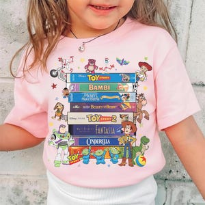 Może przedstawiać: Jasnor&oacute;żowa koszulka z nadrukiem stosu kaset VHS Disneya, z tytułami takimi jak 'Toy Story', 'Bambi' i 'Piękna i Bestia'. Kolorowe postacie z kresk&oacute;wek zdobią prz&oacute;d koszulki.