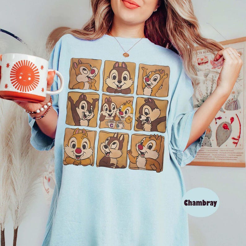 May include: Light blue t-shirt featuring a grid of Chip and Dale cartoon character illustrations in brown squares. The shirt has a relaxed fit and the word "Chambray" is printed on the bottom right.