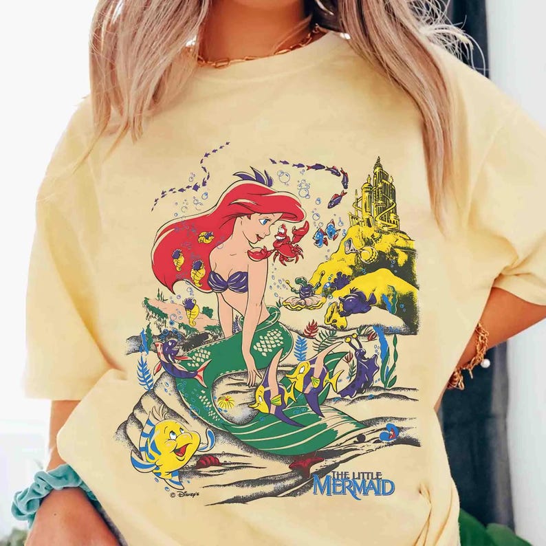 K&ouml;nnte beinhalten: Hellgelbes T-Shirt mit einer farbenfrohen Grafik von Arielle aus Disneys Der kleinen Meerjungfrau. Die Illustration zeigt Arielle mit roten Haaren, einem gr&uuml;nen Schwanz und verschiedenen Meeresbewohnern. Der Text "THE LITTLE MERMAID" steht unten.