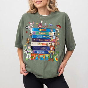 Może przedstawiać: Zielona koszulka z nadrukiem stosu kaset VHS z filmami Disneya, takimi jak "Toy Story" i "Bambi". Postacie z kresk&oacute;wek z film&oacute;w są rozrzucone wok&oacute;ł kaset. Koszulka ma luźny kr&oacute;j.