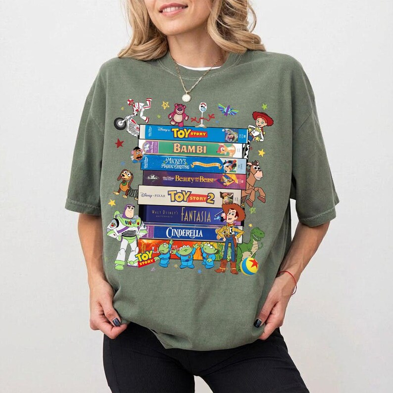 Może przedstawiać: Zielona koszulka z nadrukiem stosu kaset VHS z filmami Disneya, takimi jak "Toy Story" i "Bambi". Postacie z kresk&oacute;wek z film&oacute;w są rozrzucone wok&oacute;ł kaset. Koszulka ma luźny kr&oacute;j.