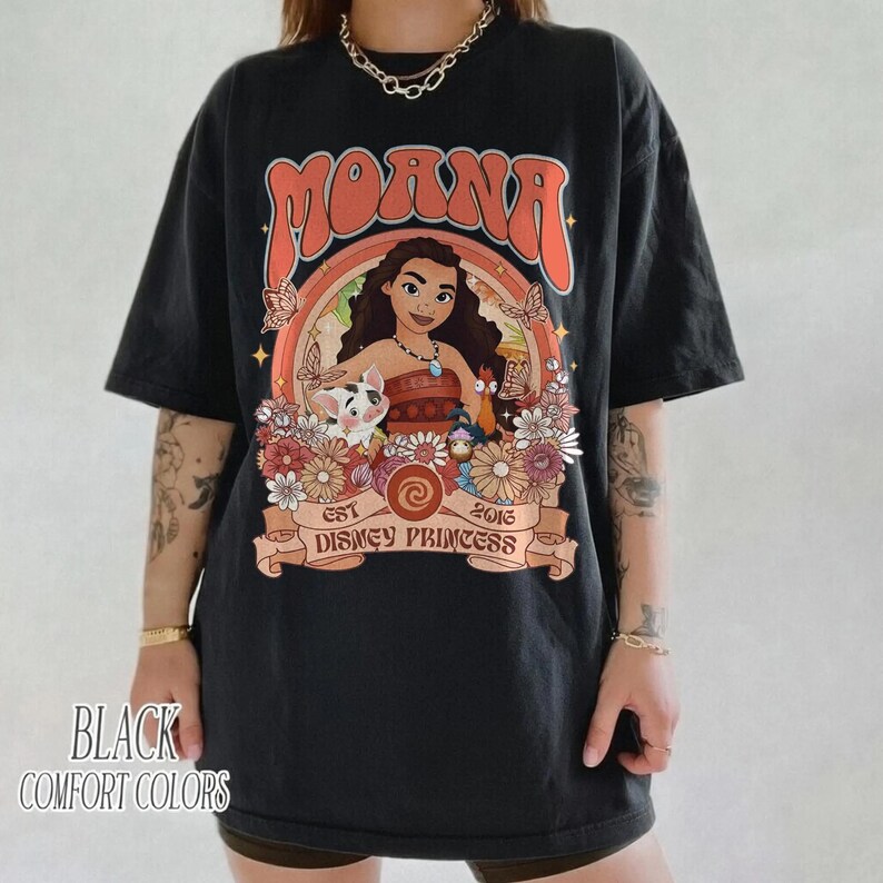 Op de afbeelding: Zwart T-shirt met een kleurrijke afbeelding van Moana, bloemen en vlinders, met het woord "Moana" in het oranje. Het shirt heeft ook de tekst "Est 2016 Disney Princess" op een banner. De woorden "Black Comfort Colors" staan linksonder.