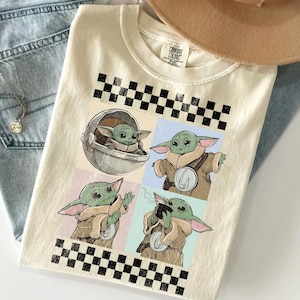 Puede incluir: Camiseta color crema con un gr&aacute;fico de cuatro paneles de un personaje alien&iacute;gena verde en varias poses. El dise&ntilde;o incluye un patr&oacute;n de tablero de ajedrez en blanco y negro y las palabras "COMFORT COLORS" en la etiqueta.