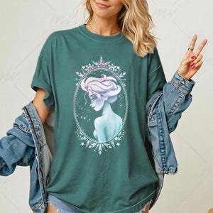 May include: Teal green t-shirt featuring a watercolor illustration of a woman with light purple hair and a blue dress inside an oval frame. The frame is decorated with snowflakes and crystals. The woman is looking over her shoulder. The shirt is paired with a denim jacket.
