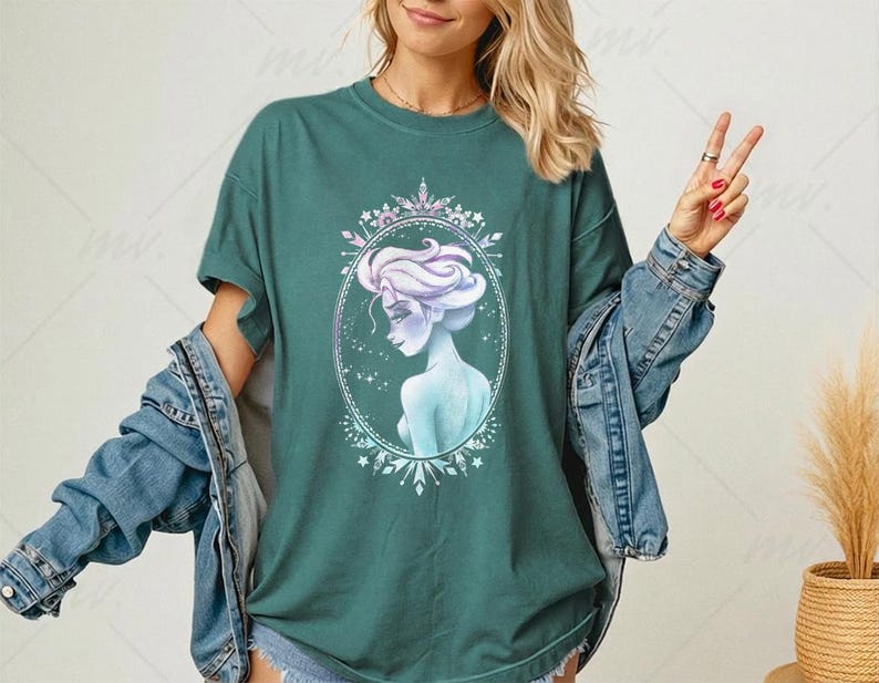 May include: Teal green t-shirt featuring a watercolor illustration of a woman with light purple hair and a blue dress inside an oval frame. The frame is decorated with snowflakes and crystals. The woman is looking over her shoulder. The shirt is paired with a denim jacket.